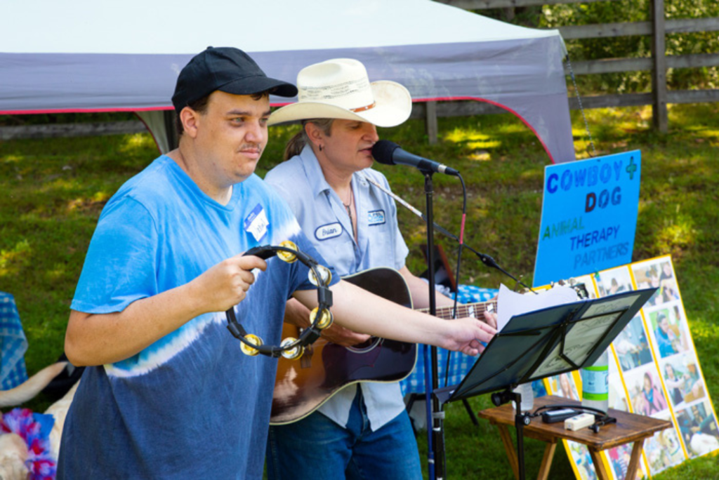 a young man playing tamborine next to man in cowboy hat playing guitar at outdoor music performance