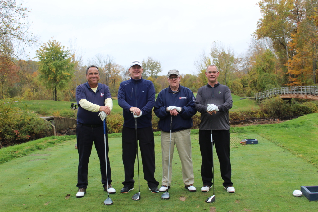 Four stand side by side on the Hollow Brook Golf course in the fall season. They each have a golf club in front of them with their hands resting on the top of the clubs.
