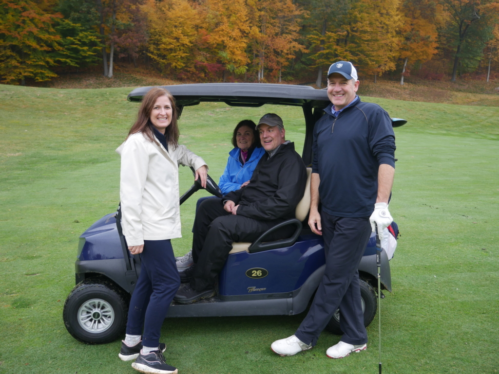 A female and male golfer stand outside a golf cart, the man has a golf club in his hand, while another couple sit inside the golf cart. The cart is parked on Hollow Brook Golf club on the green course with fall foliage in the background.