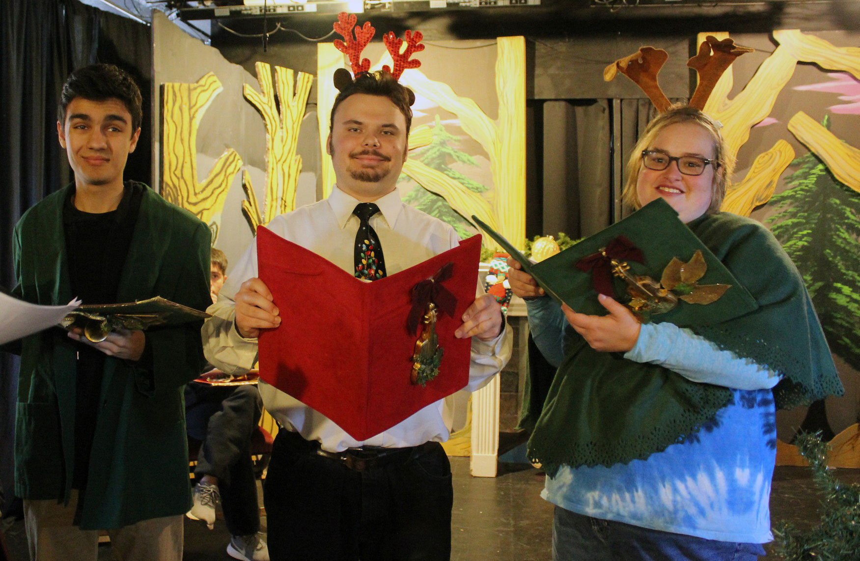 Three participants in the play "How the Grinch Stole Christmas" wear costumes and hold decorated song books while standing in a black box theater with wooded scenery.
