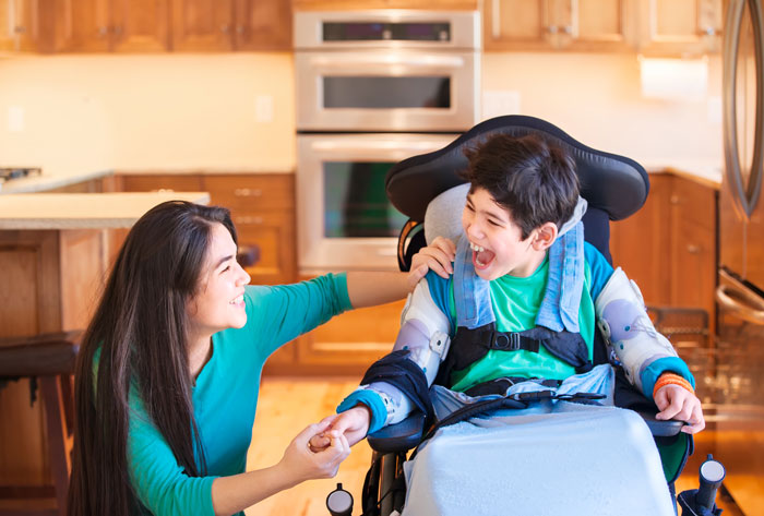 A woman kneels by a wheel chair occupied by a young person. They are holding hands and are in the kitchen.