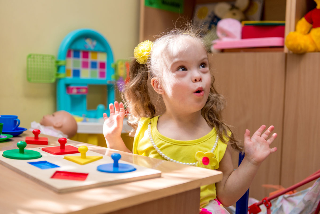 A young child with down syndrom plays with a colored shape puzzle in a bright child care setting.