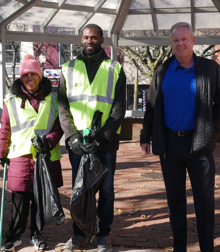 A woman and man stand in an outdoor atrium they helped to clean. They are wearing reflective work vests and hold pokers and garbage bags. A man who is there job development coach stands next to them.