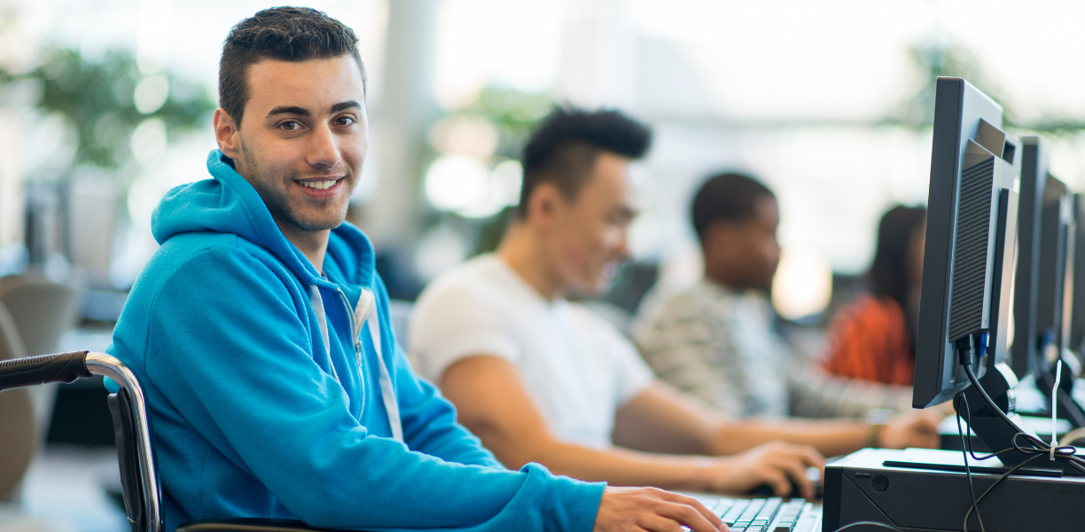 College aged man in a wheel chair sitting at a computer in a media center with other people working at computers