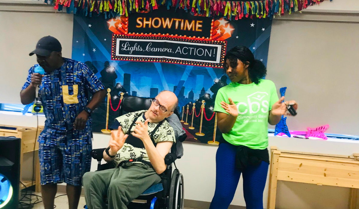 At the CBS Talent show a man wearing a baseball cap sings into a microphone while a man in a wheel chair claps his hands and a woman claps her hands to the beat.