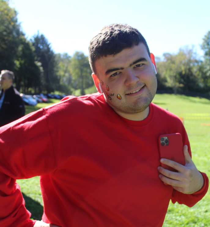 A young man dressed in red and holding a red cell phone, poses with one cheek painted outdoors during the fall festival.