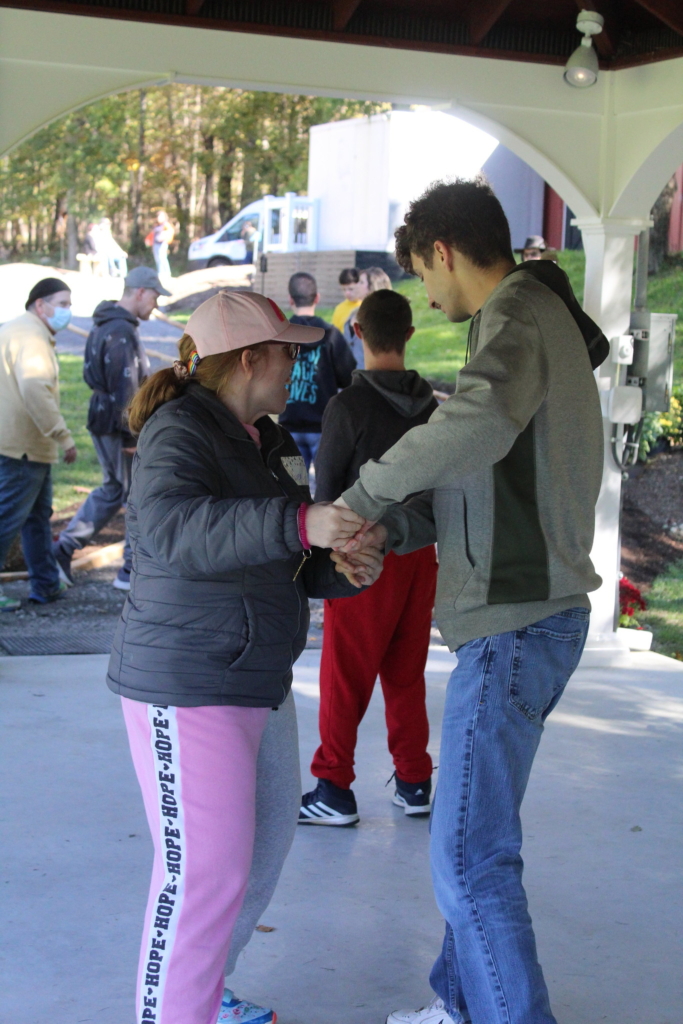 A young woman in a pink ball cap and pink sweatpants holds hands with a young man in jeans and a grey jacket as they dance under a white pavilion at Cultivating Dreams Farm