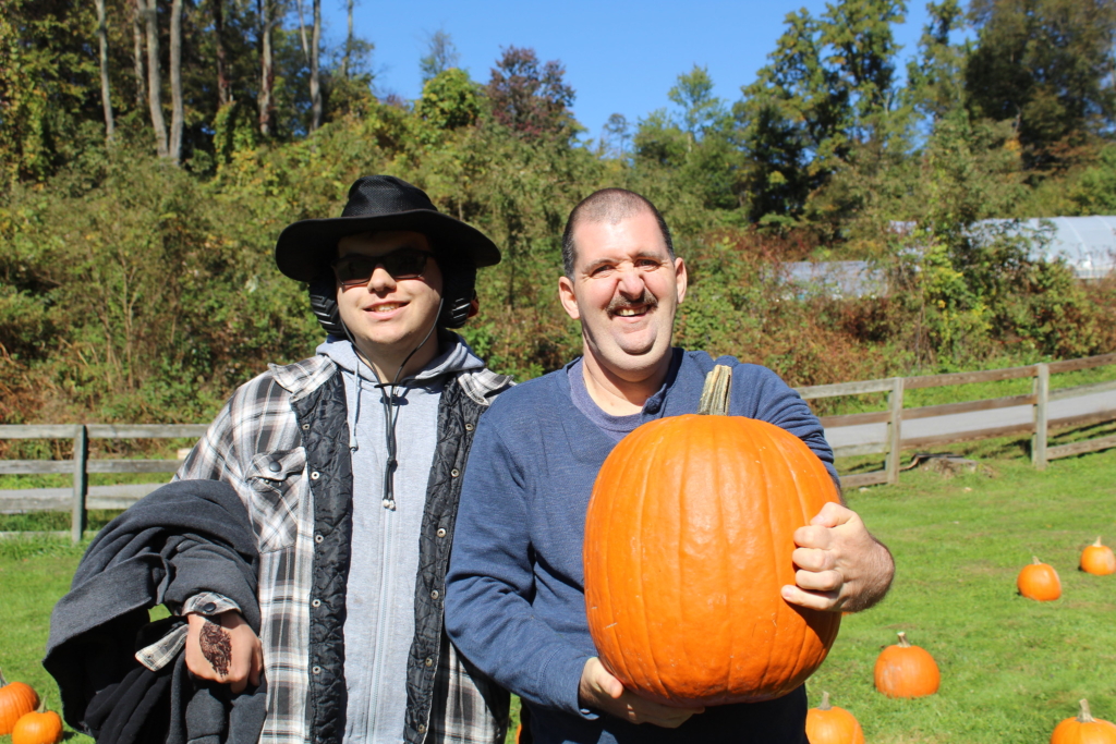 A man with a hat and flannel shirt and a man in a blue shirt holds a large orange pumpkin from the pumkin patch at Cultivating Dreams Farm
