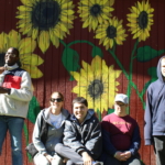 A group of people at Cultivating Dreams Farm Fall Festival stand and sit in front of a red shed with big bright yellow sunflowers painted on it.