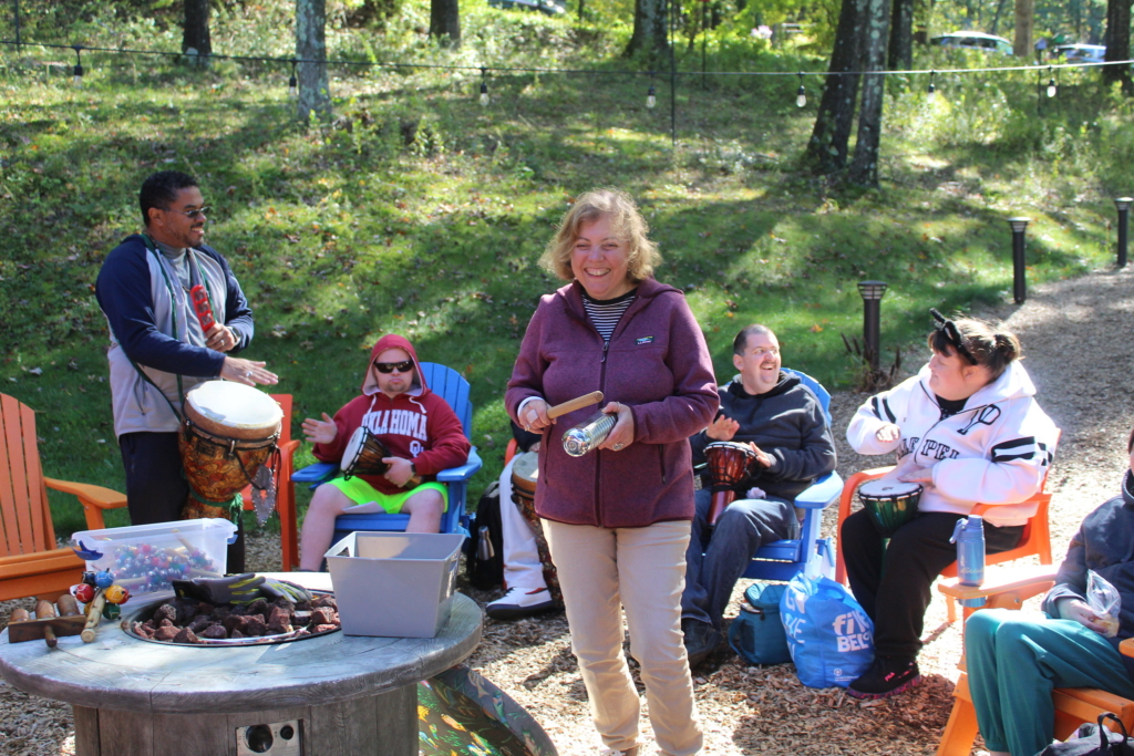 A drumming circle, led by Ernesto on the bongos, sits around a fire pit while a woman stands in front playing a rythmn instrument.