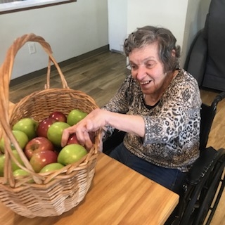 An older woman in a wheel chair sits at a table while reaching into a basket of red and green apples in a home supported by Community Based Services