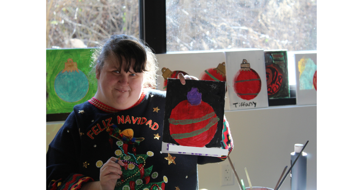 a woman in a festive holiday sweater holds up a painting of a red ornament with purple and gold design that she created at the Hudson Valley InterArts Center