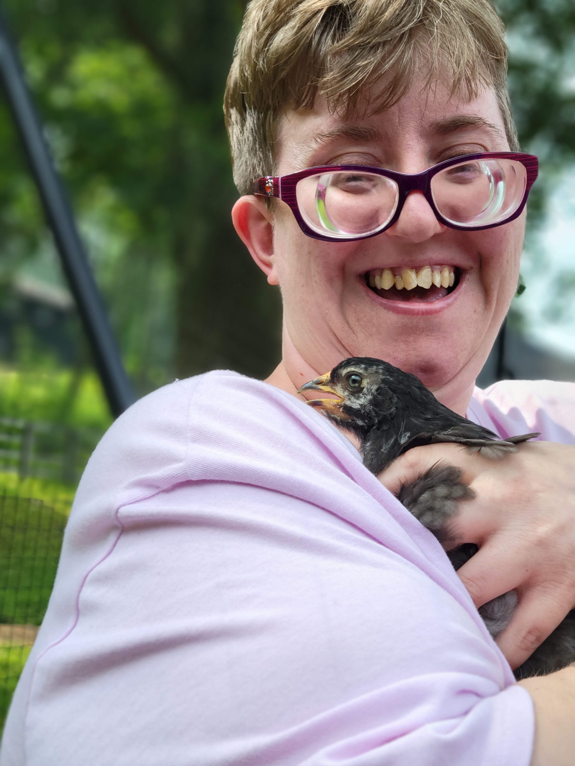 A woman with short hair and glasses holds a chicken that is a few weeks old. She is smiling.