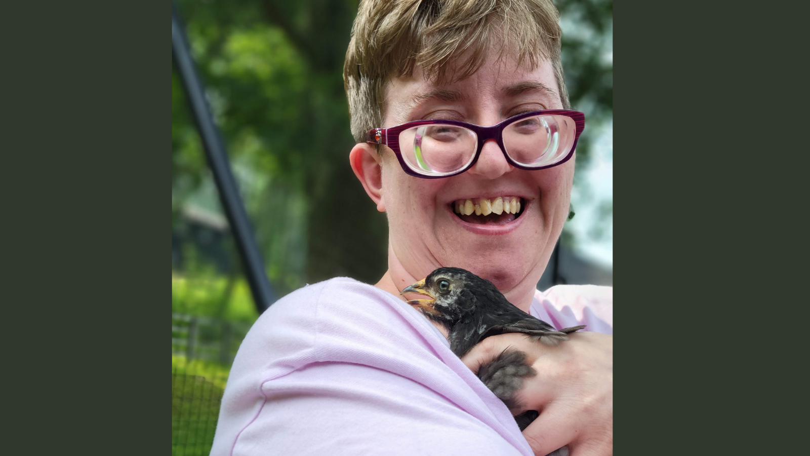 A woman with short hair and red glasses smiles as she holds a chicken that is a few weeks old.