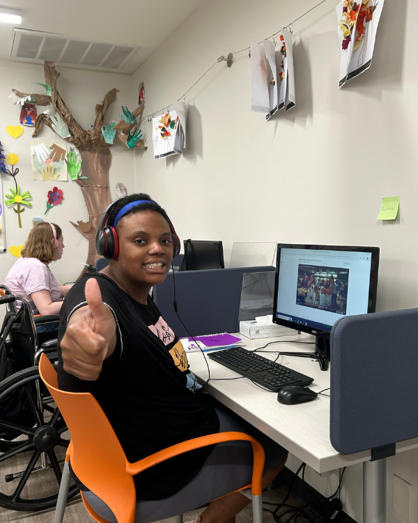 A women sits in front of a computer of the media lab at Site Based Day Hab. She is smiling, looking towards the camera with her thumb up. Another women is in the background with headphones on and she looks at her computer screen.