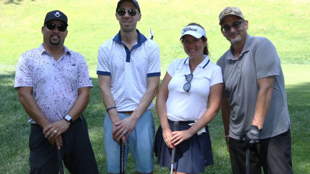 A golf foursome consisting of two men in caps and golf shirts and a woman in a visor and golf skirt pose on the greens of Hollow Brook Golf Club during the CBS Charity Golf Tournament.