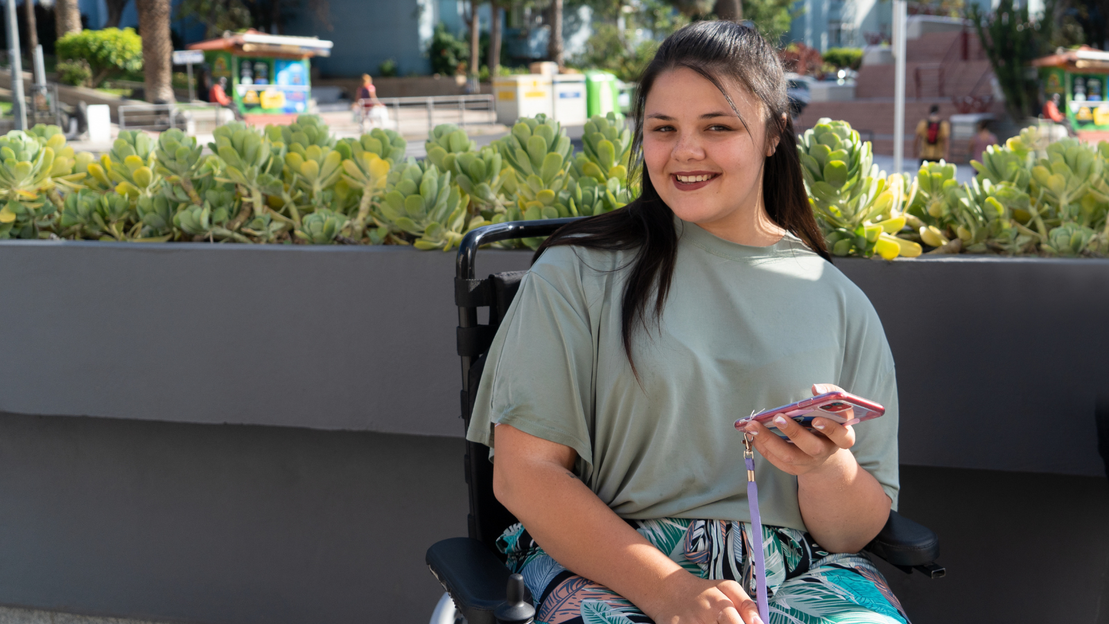 A teenage girl with long hair sits outdoors on a black chair. She holds a pink smart phone in her left hand and she is smiling.