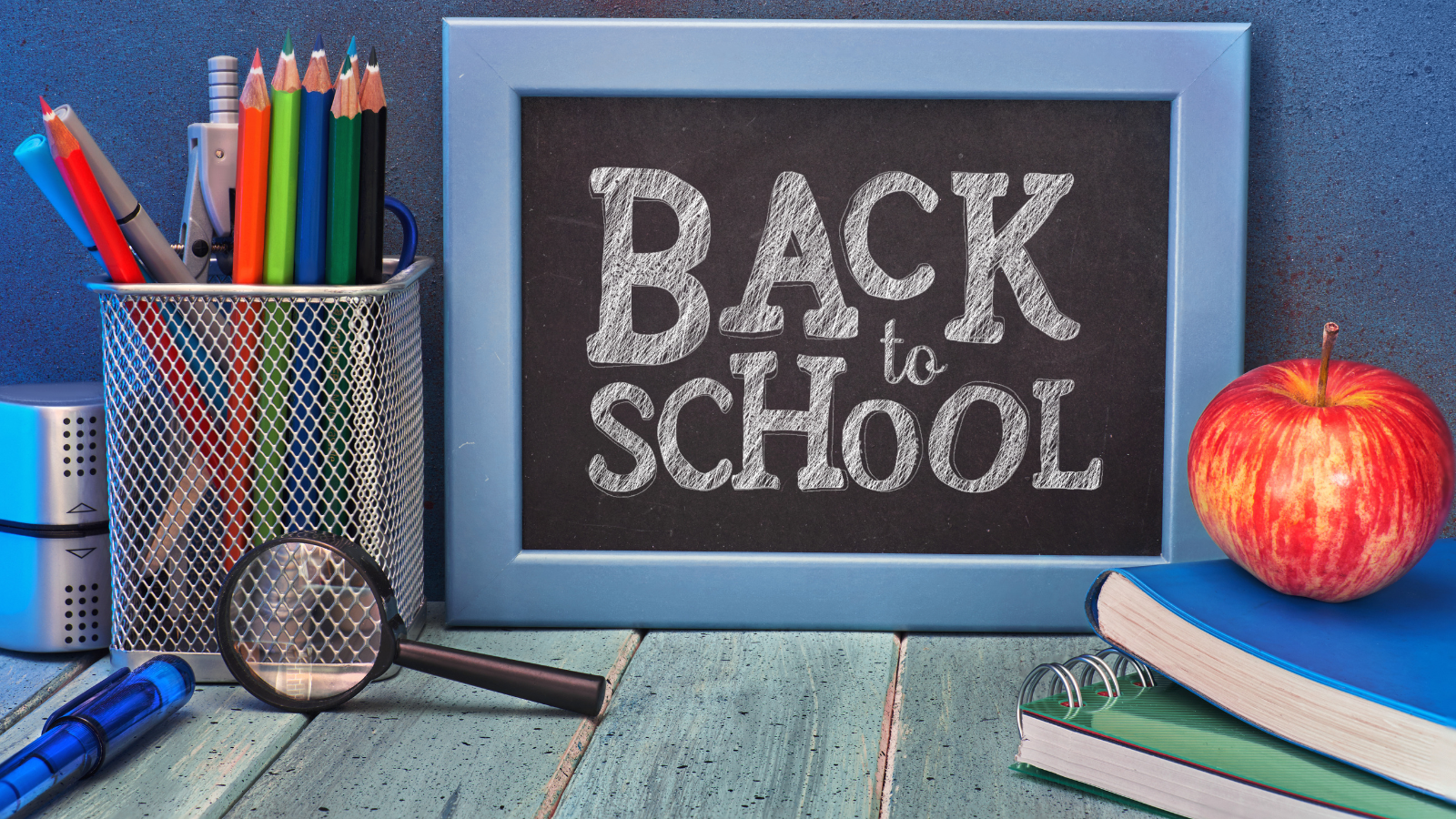 The words Back to School written in chalk on a table top sized blackboard sign sits on a desk with school supplies; books, magnifying glass, a red apple, and a container of colored pencils