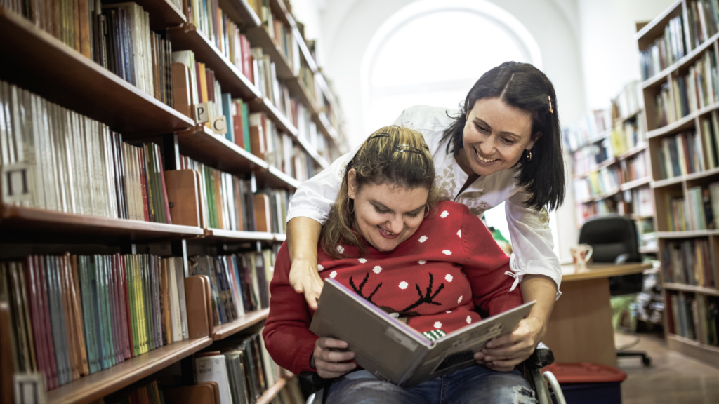 A teenage girl sits in a wheelchair while reading a book in the aisle of a library while her mom looks over her shoulder