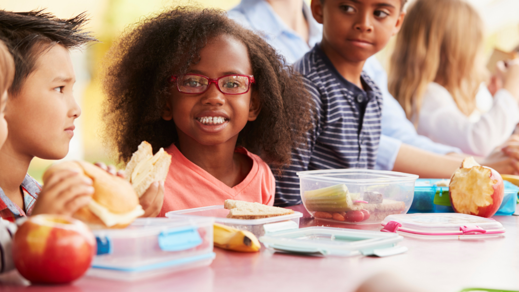 Elementary school aged children eat home made lunches around a school table.