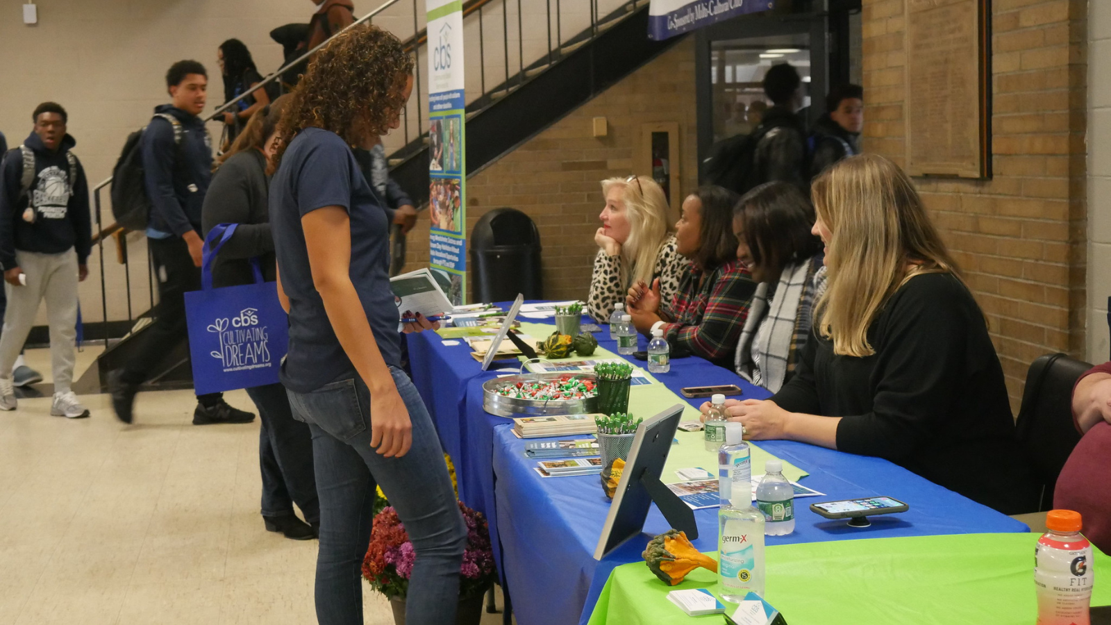 A female high school student stands at an informational table with green and blue tablecovers and information. The table is part of a fair inside a busy high school area.