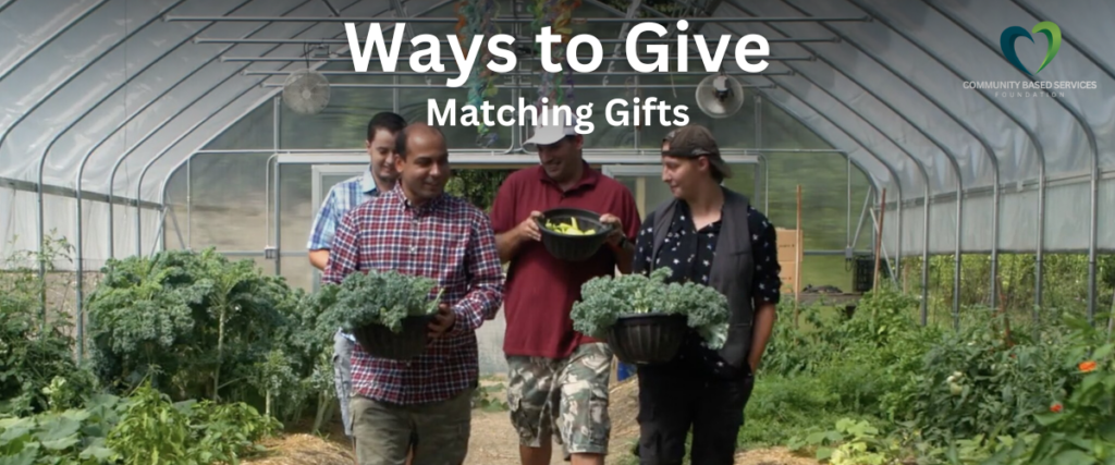 Four people walk through a greenhouse lush with growing plants as they carry baskets of produce. They are looking towards each other and smiling.