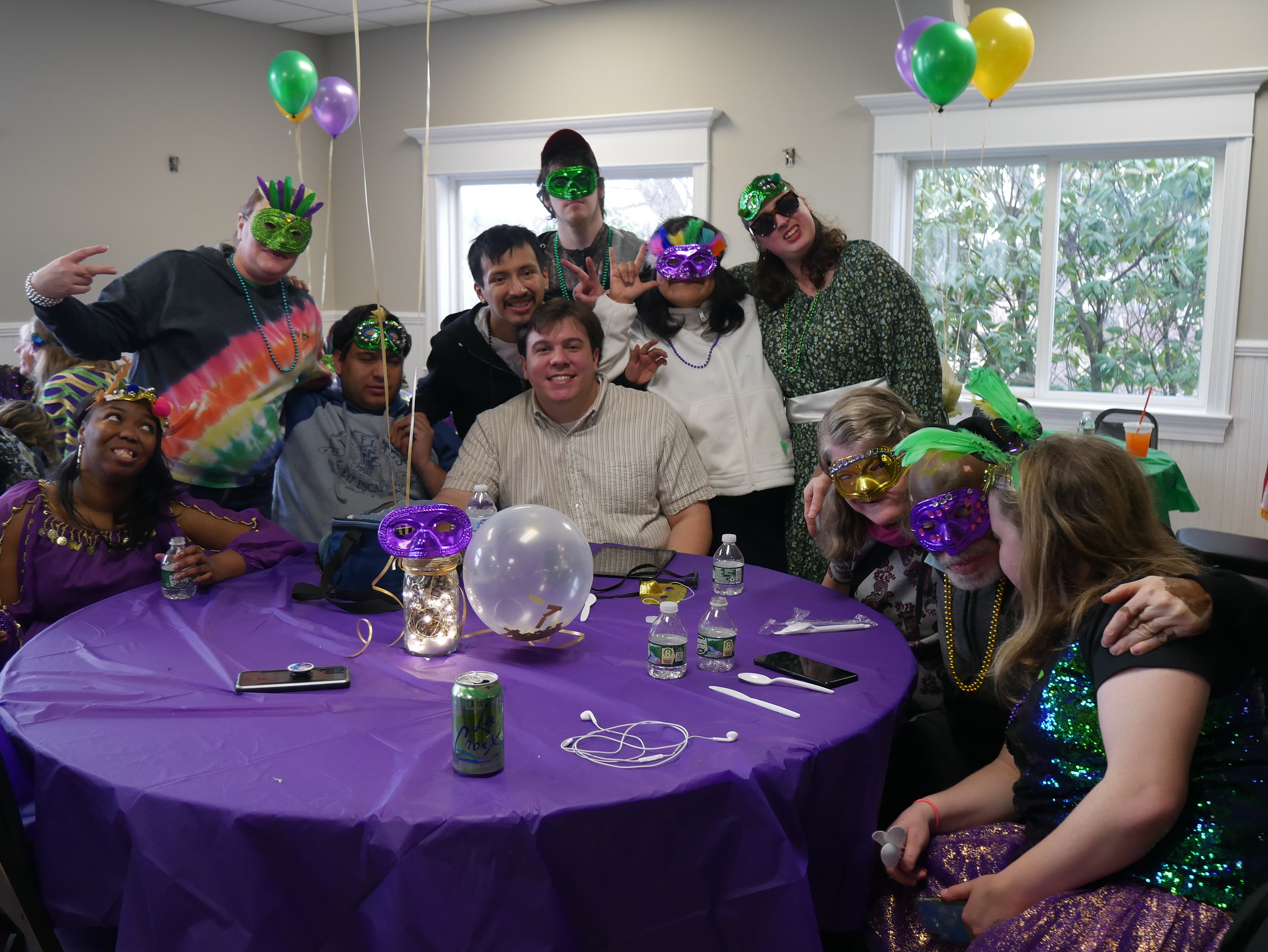 A group of friends supported by CBS adorned in mardi gras masks sit around a purple clothed table, smiling and with their arms around each other.
