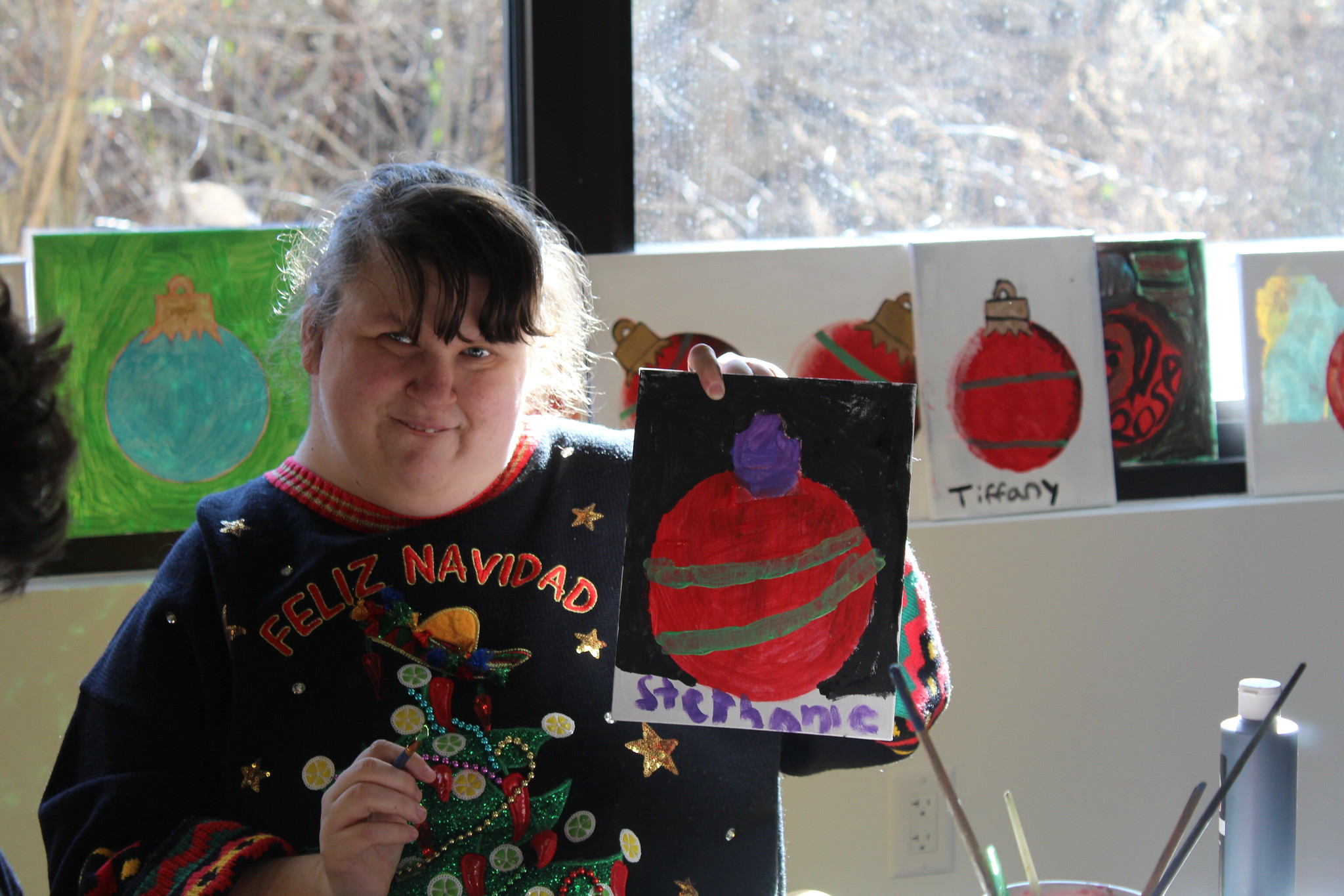 A young women at the soft opening of InterArts in December 2023 wears a festive holiday sweater and proudly holds up her painting of an ornament.