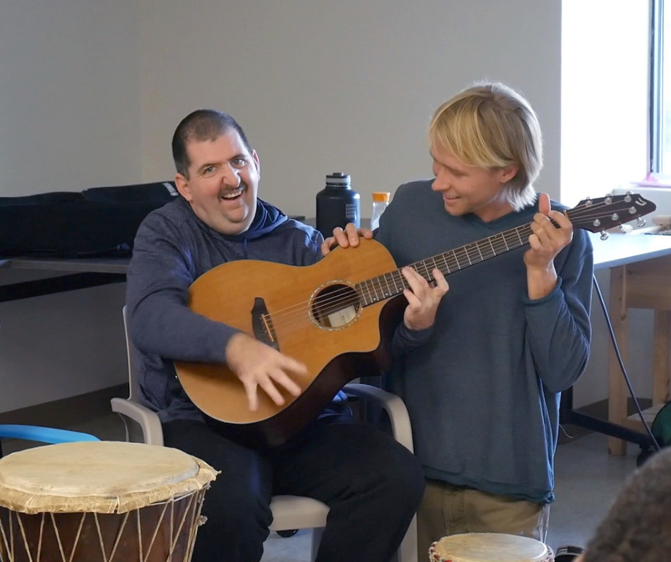 A man smiles while strumming an acoustic guitar. His music instructor looks on while helping him find the chords at a day hab program at InterArts in Patterson, NY.