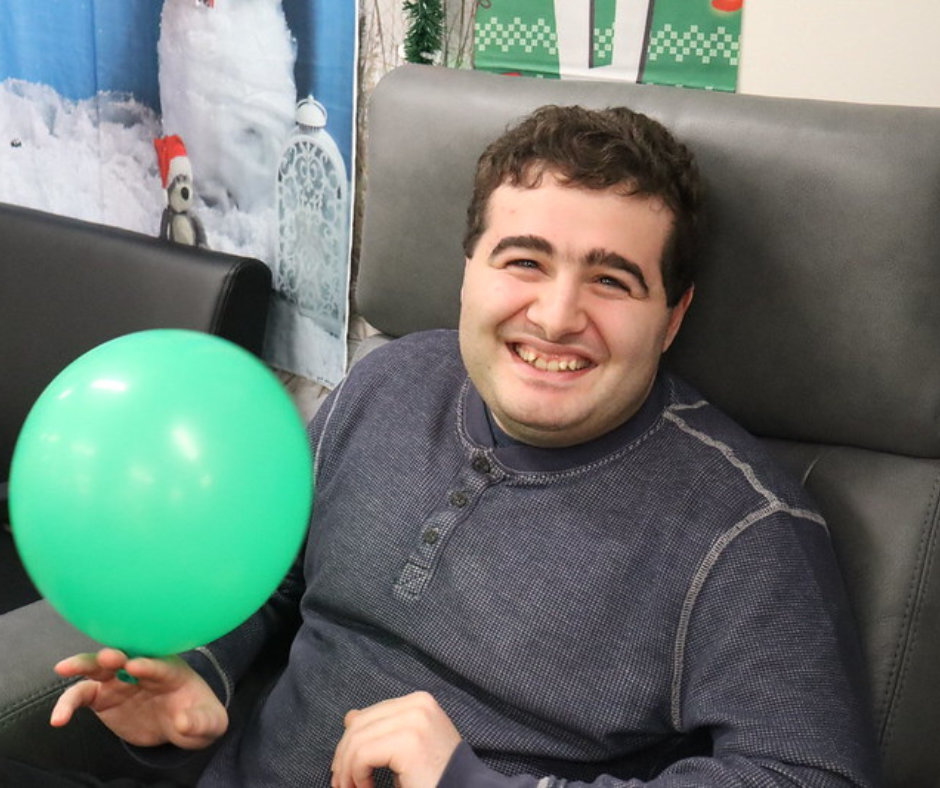 A man in the site based day hab program smiles as he relaxes in a massage chair while holding a party balloon during a holiday luncheon.