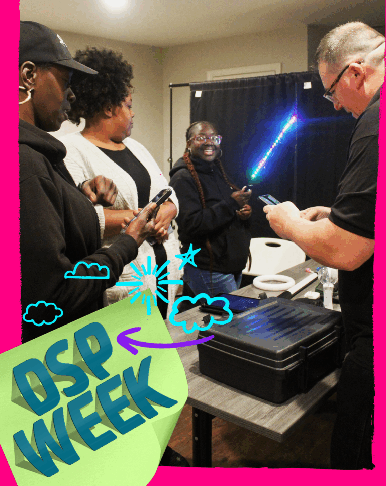 A group of DSPs engage in a photographic light painting class under the leadership of InterArts instructor Jonathan Harrison. They are gathered around their cell phone cameras and one woman uses a light wand in the background.