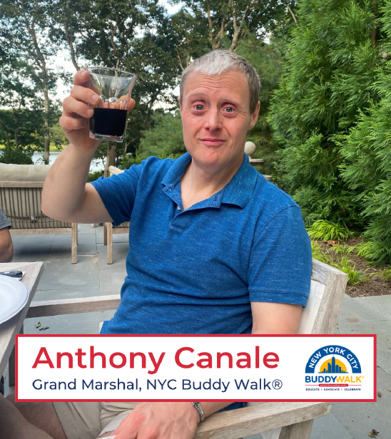 Anthony Canale seated outside at a table and holding up a glass as in a toast. He is wearing a blue polo style shirt and trees and shrubbery are in the background.