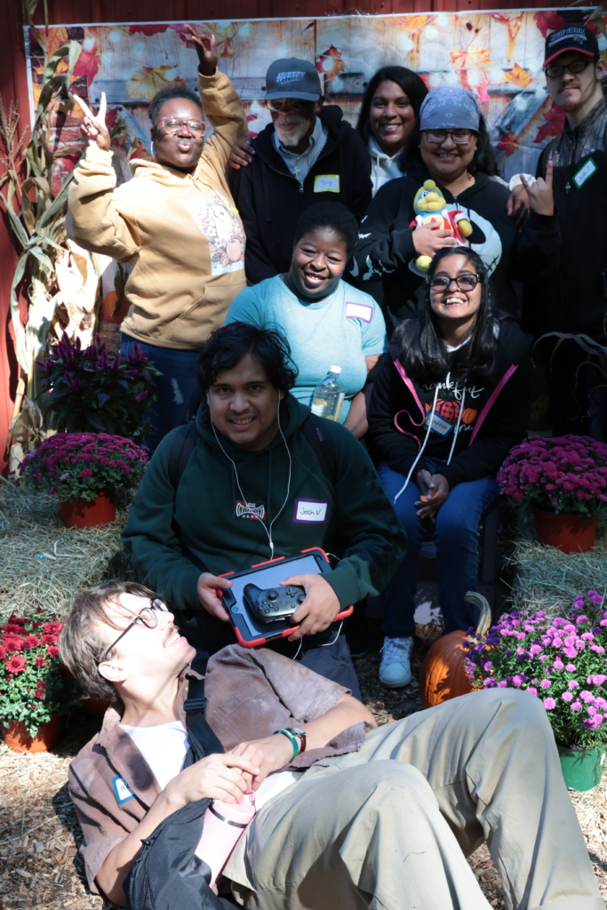 A day habilitation group gathers in front of a fall backdrop with pots of mum flowers and cornstalks displaying smiles and silliness as they have their photo professionally taken.