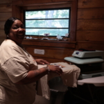 A woman smiles as she works a tee shirt press in a rustic barn