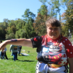 A woman smiles as she holds out her arm to show off a temporary tattoo. Behind her people, trees and pumpkins dot the landscape.