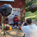 A young woman sits on a hay bale in front of a barn door back drop surrounded by hay bales, potted mums and cornstalks. She is posing for the photographer who is about to take the photo. Professional photographer equipment and lights are in the scene.