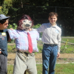 Two men stand on either side of scarecrow they just created. The scarecrow wears a wig, bright red tie, khaki pants and a plaid shirt. They stand in front of a garden.