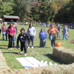 A woman is poised to throw a bowling ball down the hay bale lane in an attempt to knock down the pins. Her group and staff members cheer her on.