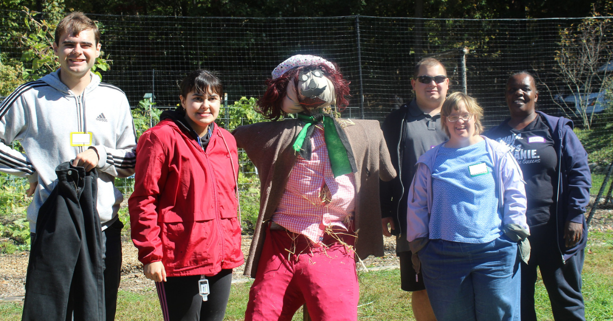 A CBS day hab group stands with a whimsically dressed scarecrow they created together at Cultivating Dreams Farm during Fall Festival. The outdoor garden appears in the background.