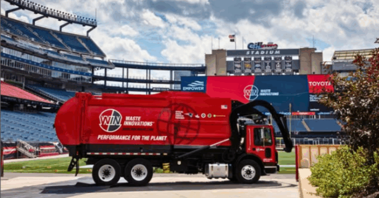 Large red waste removal truck with Win Waste Innovations logo. The vehicle it parked at Gillette Stadium and the stands rise behind the truck.