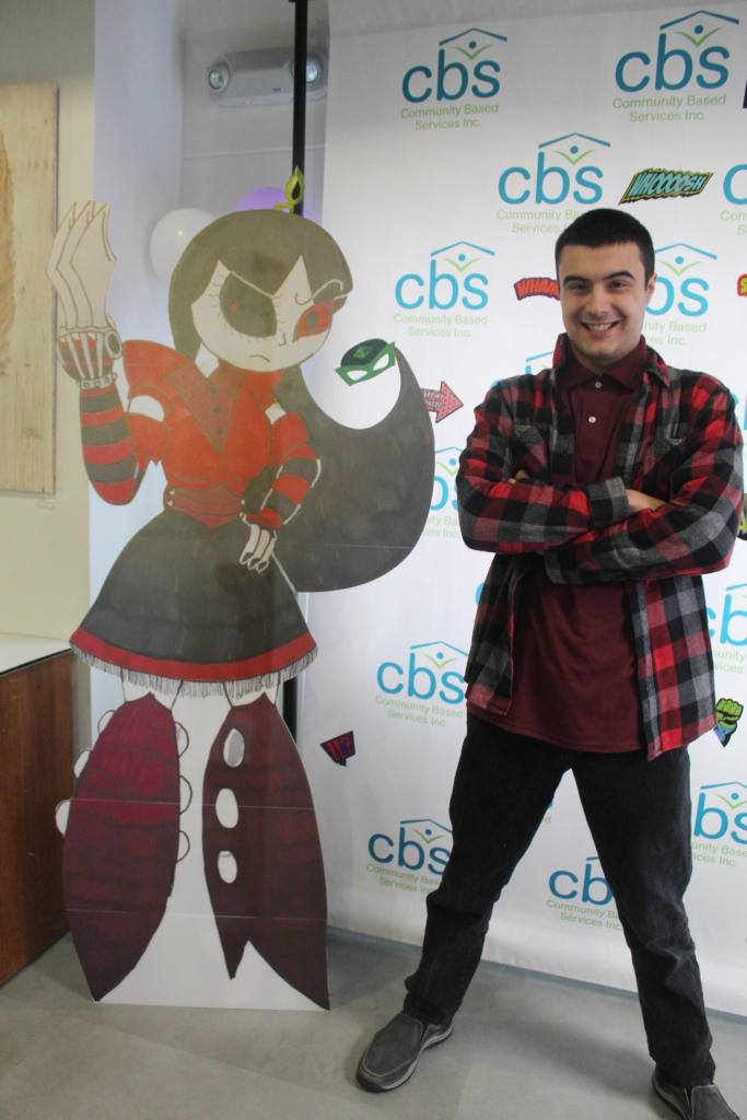 Creator Alex stands with arms crossed, smiling next to his life sized character stand, Belladonna.