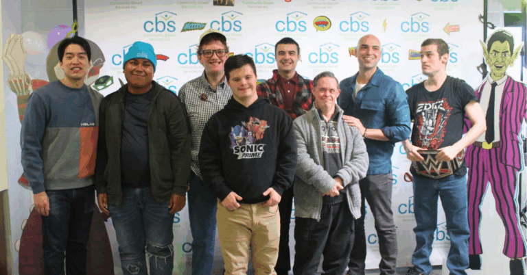 Six men in the CBS group day hab and creators of the Zero to Hero comic book stand with Artist Brian Chan and Group Day Hab Manager Frank DiMaggio. They group stands in front of a CBS logo branded back drop with comic book speech bubbles. Two character stands from the book are on either side of the group.