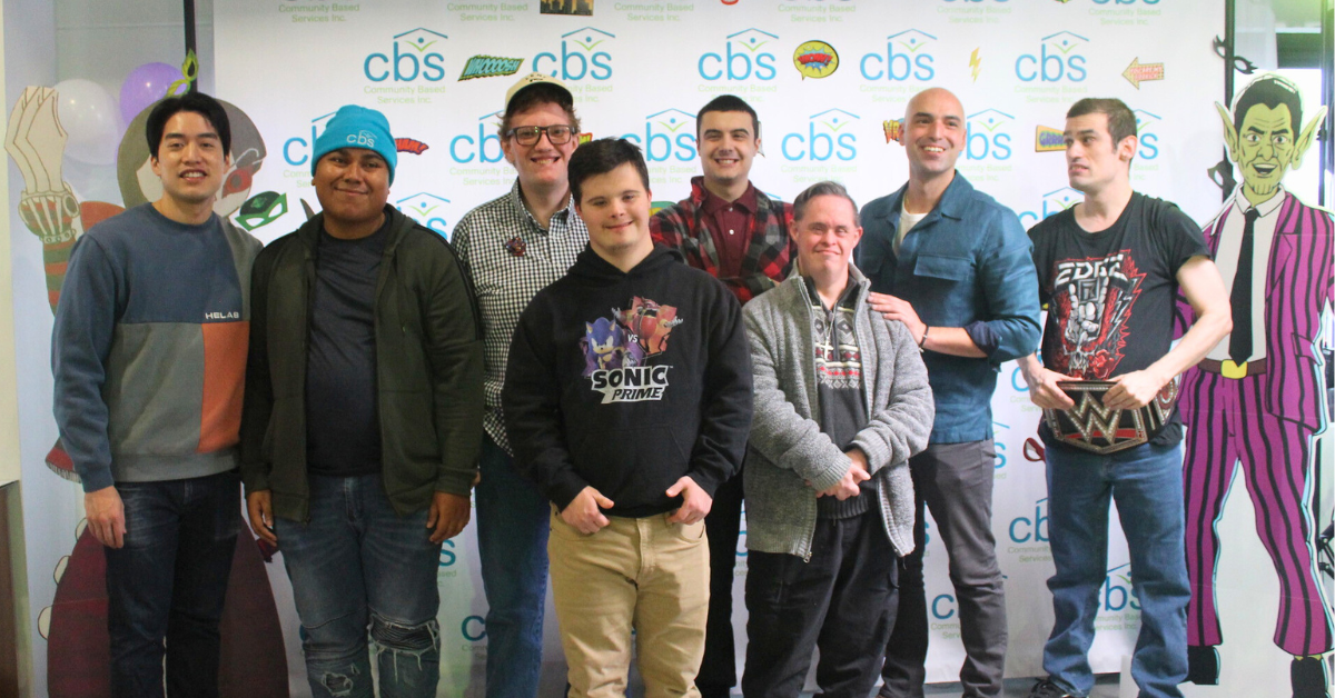 Six men in the CBS group day hab and creators of the Zero to Hero comic book stand with Artist Brian Chan and Group Day Hab Manager Frank DiMaggio. They group stands in front of a CBS logo branded back drop with comic book speech bubbles. Two character stands from the book are on either side of the group.