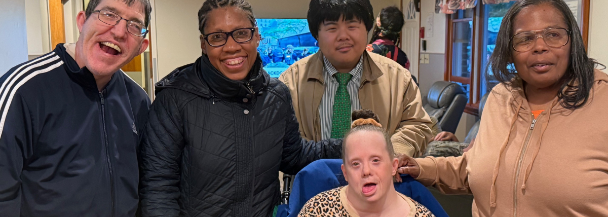 Friends residing in CBS homes visit one another. They stand with another resident who is in a wheel chair and everyone is smiling. A direct support professional stands with them.