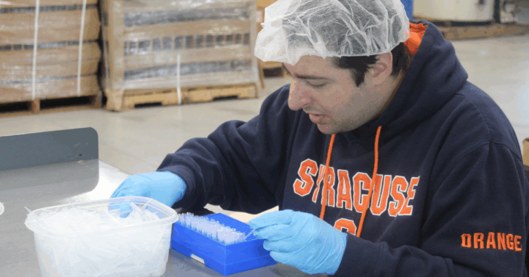 A worker wearing a hair net and blue gloves packs plastic pipettes into a special container in the manufacturing floor of Vista Lab. He is surrounded by palettes and machinery.