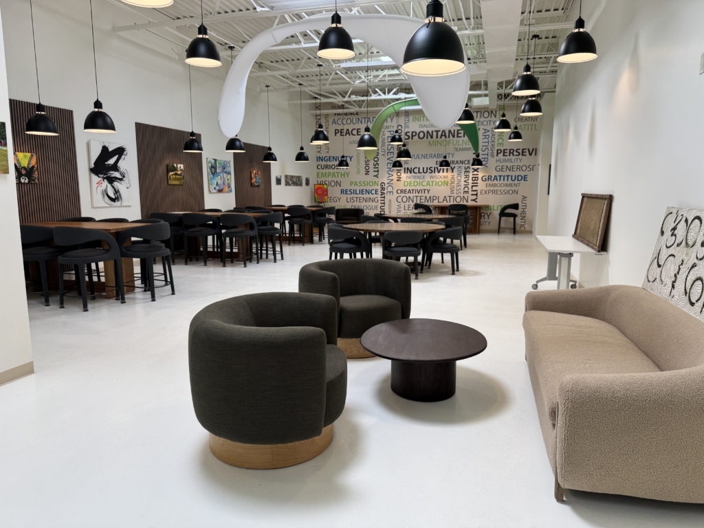 The main interior of the Impact Center for Learning with a white floor and walls, art, and word mural. Comfortable modern couch, chairs and tables offer the comfort of home in a work environment. Large urban metal lamps hang down from the high ceiling.