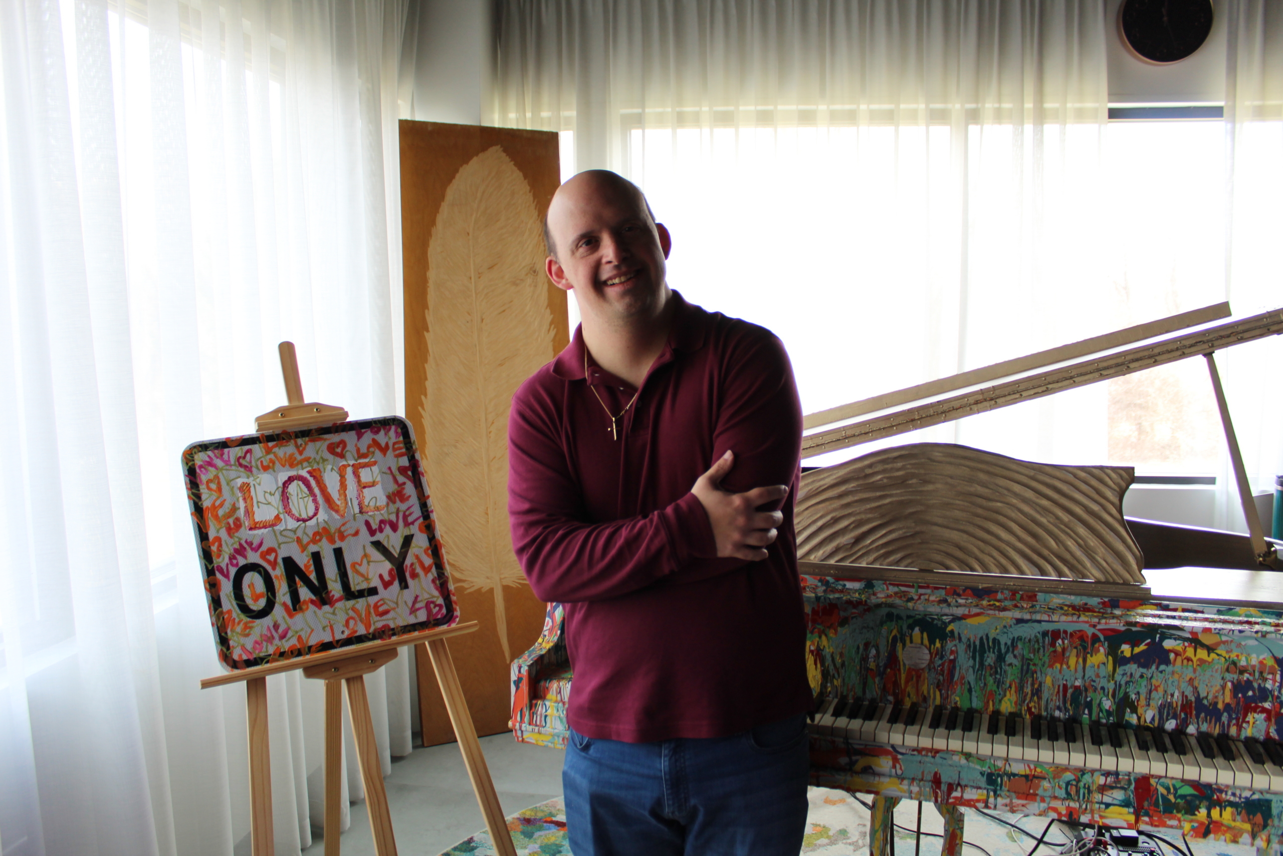 Brandon T. stands by a painted piano and a sign that says "Love Only." He leans agains the piano in a maroon collared shirt, jeans and a gold cross necklace with arms loosely crossed.