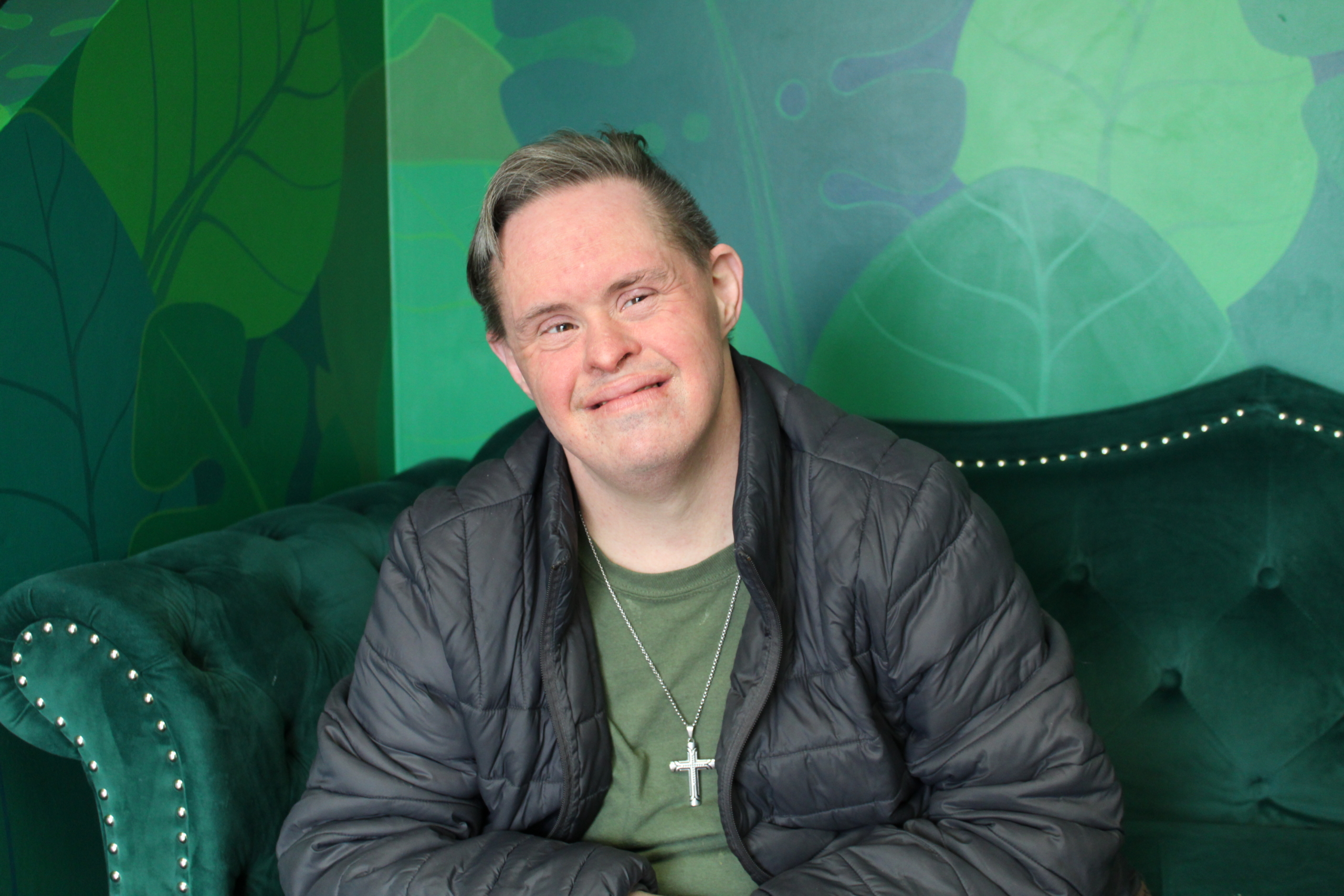 Freddy A. sits on a green velvet upholstered couch in front of a mural of green leaves. He wears a green tee shirt, grey jacket and a large silver cross.