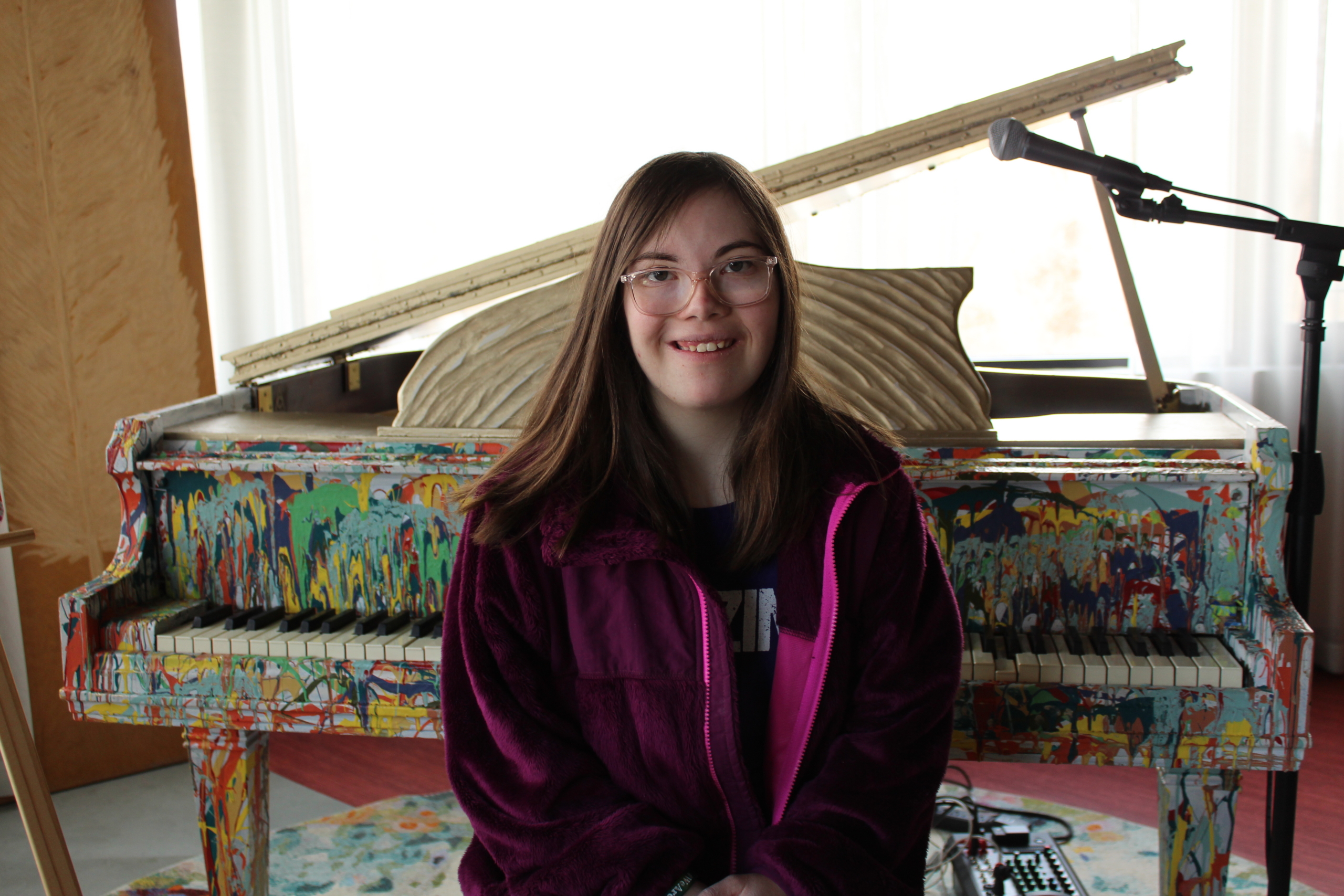 Haley sits on a piano bench in front of the splash painted piano at InterArts.