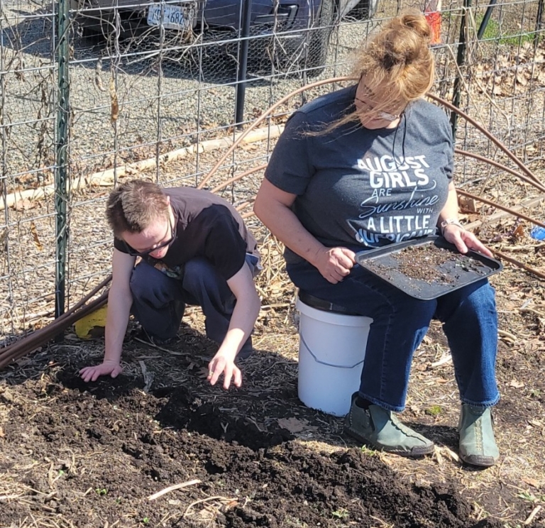 A young man learns agricultural skills under the guidance of Cultivating Dreams farmer Olimpia Bernard. They work in the soil of the outdoor garden under the early spring light.